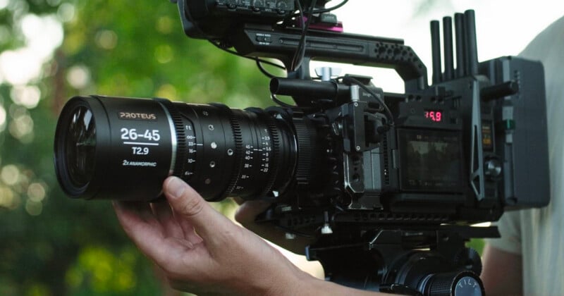 Close-up of a person holding a professional camera with a large Proteus 26-45mm T2.9 lens. The camera is mounted on a rig, with detailed controls and a small monitor screen visible. Green foliage is blurred in the background.