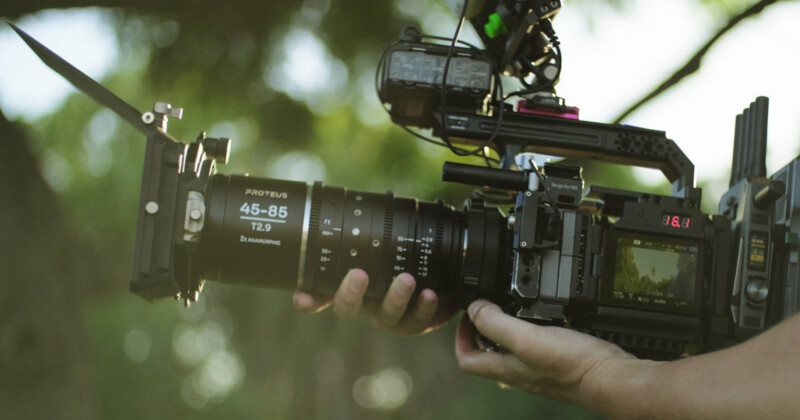 Close-up of a person holding a professional camera outdoors. The camera has a large lens with "Proteus 45-85 T2.9" written on it, and various accessories attached. The background is blurred green foliage.
