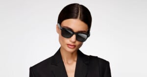 A person with dark hair wears black sunglasses and a black blazer, posing against a plain white background.