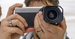 A person holds a Realme smartphone with an attached lens, resembling a high-end camera setup. The phone and lens are held with both hands, highlighting silver rings on the fingers. The background is blurred.