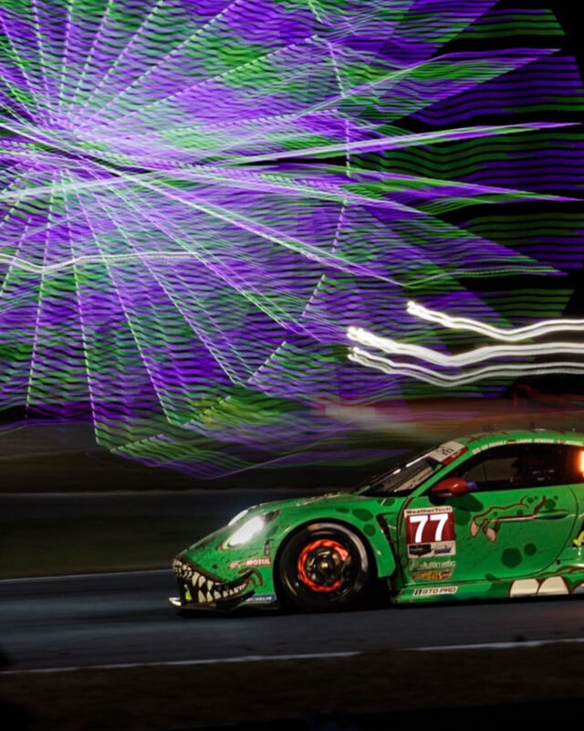 A green race car with number 77 drives on a track at night. In the background, a purple and green ferris wheel creates a colorful light blur, adding dynamic motion to the scene. The car features a crocodile theme design.
