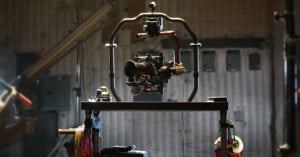 A professional video camera mounted on a Ronin stabilizer sits on a tripod in a dimly lit studio. Equipment and cables are visible in the background, giving a sense of a busy production environment.