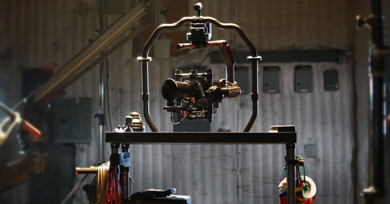 A professional video camera mounted on a Ronin stabilizer sits on a tripod in a dimly lit studio. Equipment and cables are visible in the background, giving a sense of a busy production environment.