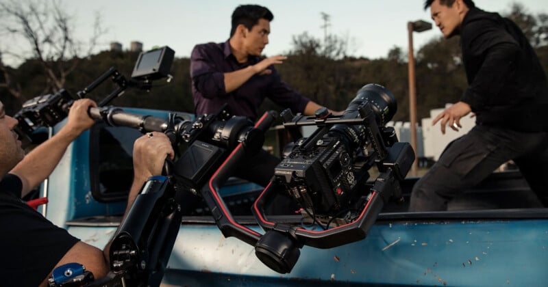 A film crew captures an action scene with two actors engaging in a fight on the back of a moving truck. The camera is mounted on a stabilizing rig. Trees and a building are in the background.