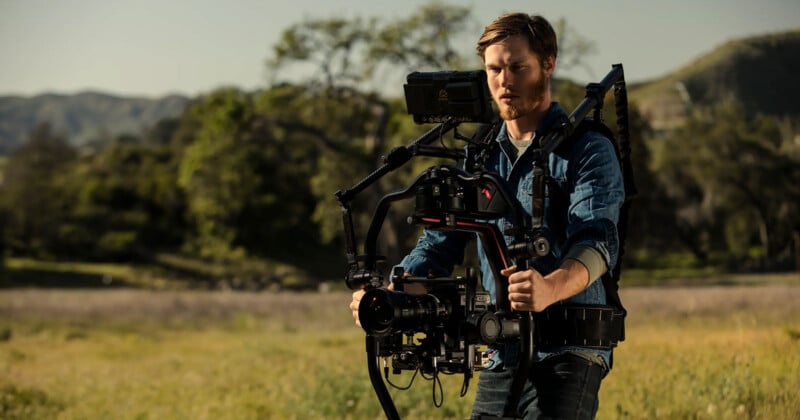 A person outdoors operates a professional camera rig with stabilizing gear. The background features a grassy field and trees under a clear sky. The individual is wearing a denim jacket and is focused on handling the equipment.