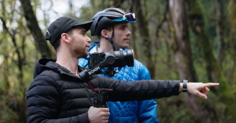 Two men are outdoors in a wooded area. The man on the left holds a camera on a stabilizer and points forward, while the other man in a blue jacket and helmet looks in the same direction. Both appear focused on something off-camera.