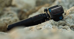 A black action camera with a grip attachment rests on light-colored rocks. The camera has a wide lens and a small screen, set against a blurred rocky background.