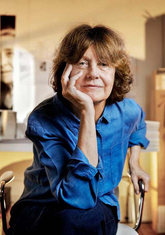 Person with shoulder-length brown hair, wearing a blue shirt, sitting and resting their head on their hand, smiling slightly. There is a blurred background with books and a partially visible black-and-white photo.