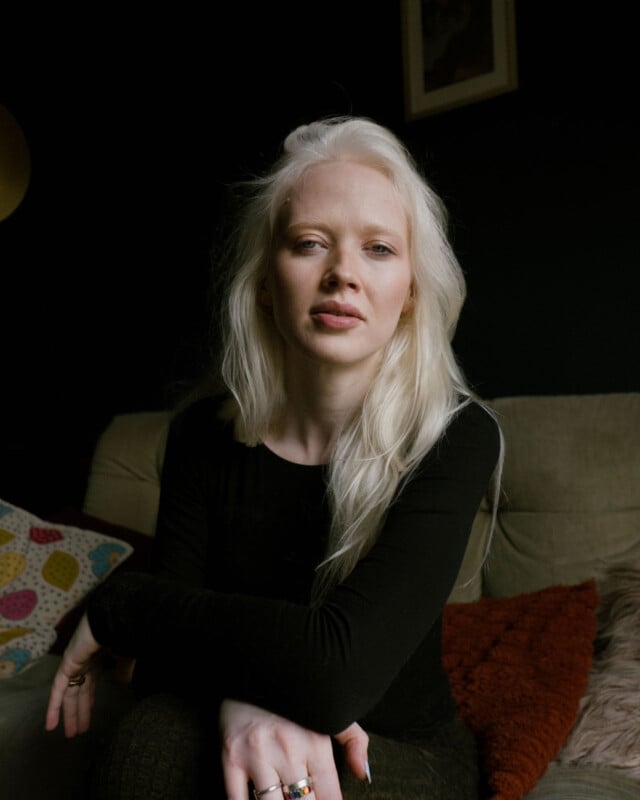 A woman with long, blonde hair sits on a couch with her arms crossed. She wears a black top and rings on her fingers. The background is dimly lit, with colorful pillows and a framed picture on a dark wall.