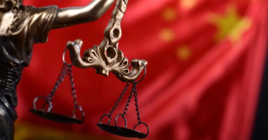 Close-up of a bronze Lady Justice statue holding scales, with the red flag of China featuring yellow stars blurred in the background.