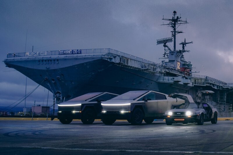 Two futuristic vehicles parked beside a large battleship at a harbor. The vehicles have sleek designs with LED lights. The battleship is gray and massive, with visible radar equipment and communication towers on deck. The sky is overcast.
