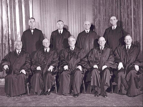 A historical black-and-white photograph of nine men in judicial robes. They are posed in two rows, with five seated in front and four standing behind them. The setting appears formal, possibly a courtroom or government building.