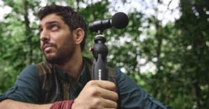 A man with a beard and a blue shirt sits outdoors in a forest, holding a small microphone attached to a tripod. He is looking to the side, and there is lush green foliage in the background.