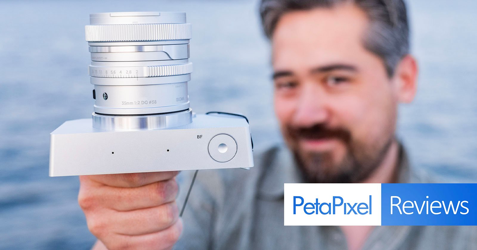 A person holding a silver camera toward the viewer, with an out-of-focus background of water. The photo features the text "PetaPixel Reviews" on the bottom right.