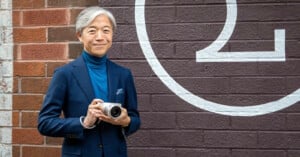 A smiling man with gray hair holds a camera, standing against a brick wall painted with a large, partial number two. He is wearing a blue turtleneck sweater and a navy suit.