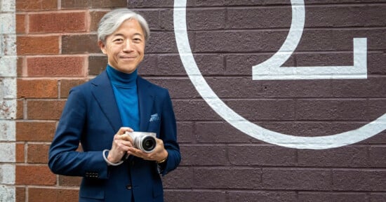 A smiling man with gray hair holds a camera, standing against a brick wall painted with a large, partial number two. He is wearing a blue turtleneck sweater and a navy suit.