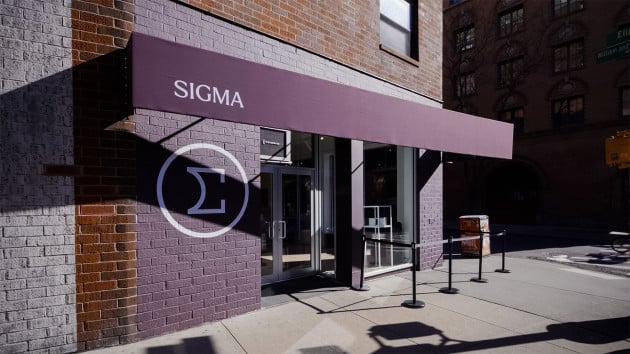 Storefront with a maroon awning displaying "SIGMA," featuring double glass doors and a Greek letter sigma symbol on a brick wall. Metal stanchions line the entrance, and a sunny urban street is visible in the background.