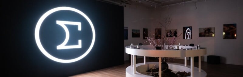 Exhibit room with a large, glowing Sigma symbol on a dark wall. In the foreground, a circular white display table with plants and small objects. Framed photos are hung on the right wall.