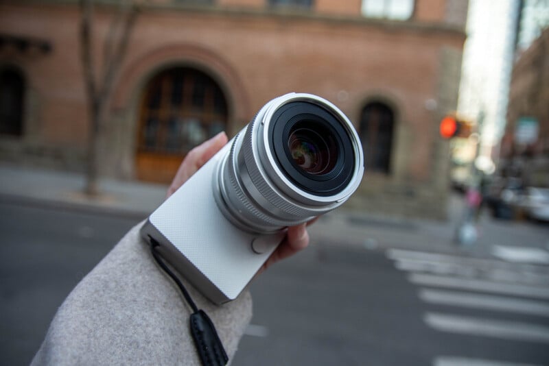 Close-up of a person holding a camera with a large lens, focusing on the lens. The background shows an urban street scene with a brick building, blurred pedestrians, and traffic lights.