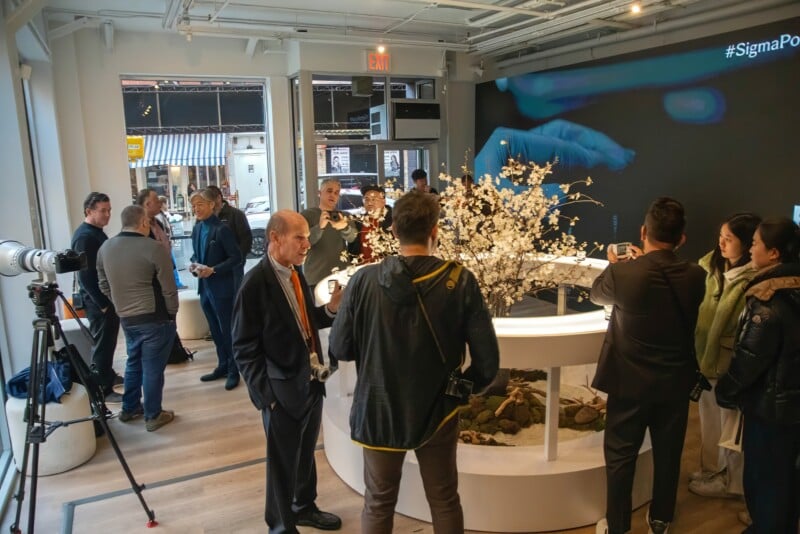 A group of people gather in a modern showroom with large windows. They are engaged in conversation and photographing a central display featuring a floral arrangement. A large screen displays an image of a hand. A camera setup is visible on the left.