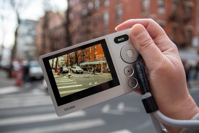 A hand holds a digital camera, capturing a busy urban street scene with pedestrians and cars. The camera display shows the scene in focus, highlighting brick buildings and a crosswalk.