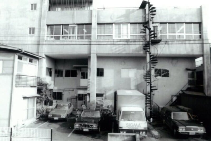 Black and white photo of a building with a narrow spiral staircase on the exterior. Below are several parked cars and a truck labeled "SIGMA." The architecture is utilitarian, with large windows on the upper floor.