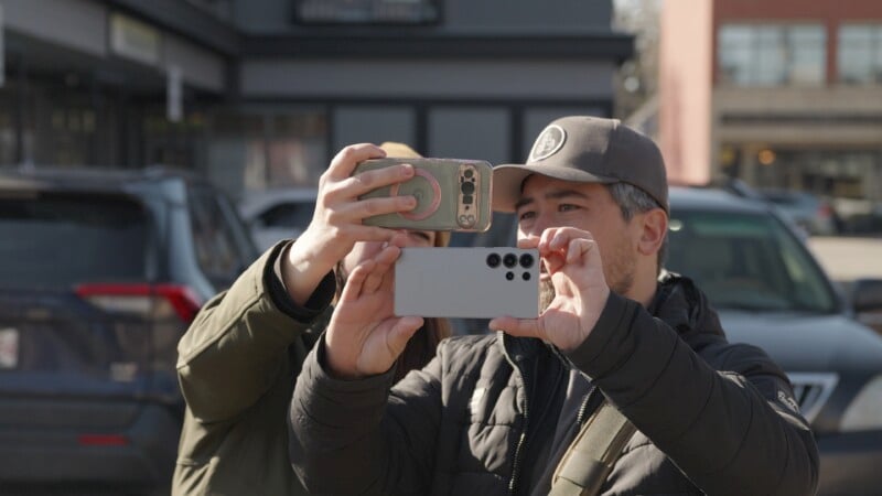 Two people are outdoors, taking photos or videos with their smartphones. They are focused on their screens, with one holding a gray phone and the other holding a phone with a case. They are dressed warmly and standing in front of buildings and parked cars.