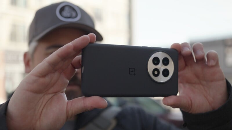 A person in a gray cap holds a smartphone horizontally, capturing a photo. The phone has a black case and a large circular camera module with three lenses. The background is blurred.