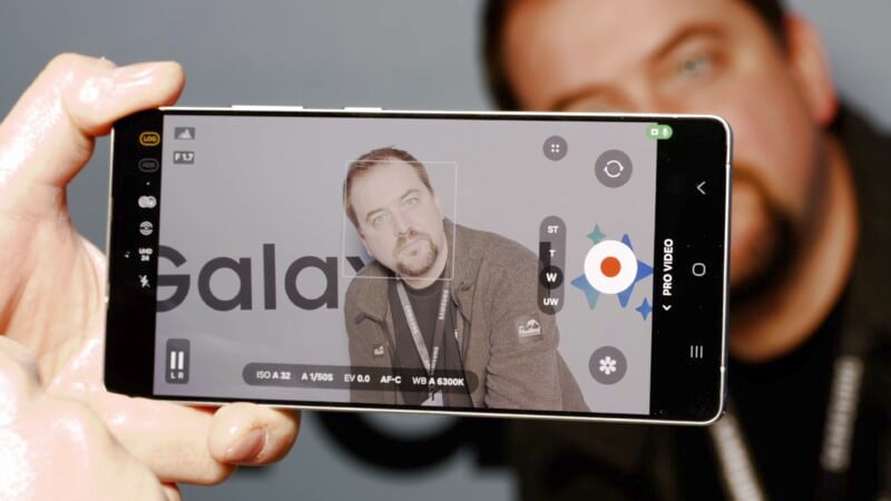 A smartphone camera focuses on a person standing against a backdrop with the word "Galaxy." The screen displays various settings and a face detection square around the person, who is wearing a lanyard and a dark jacket.