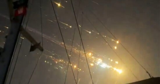 Bright streaks of light and sparks trail across a night sky, creating a dramatic scene. Dark structural outlines are visible in the foreground, possibly of a ship's mast or similar framework, illuminated by the fiery spectacle.