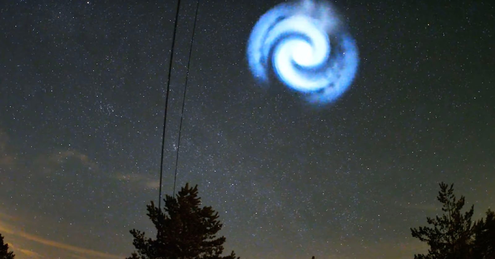 A night sky with a swirling spiral of light among stars. In the foreground, silhouettes of trees and power lines are visible. The spiral resembles a galaxy or vortex with a bright white center and blue edges.