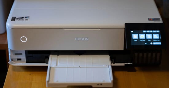 A white Epson printer on a wooden surface, featuring an extended paper tray. The printer has a small digital display screen on the right, showing icons for copy, print photos, and various prints.