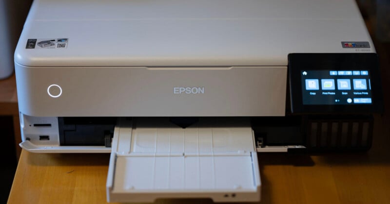 A white Epson printer on a wooden surface, featuring an extended paper tray. The printer has a small digital display screen on the right, showing icons for copy, print photos, and various prints.