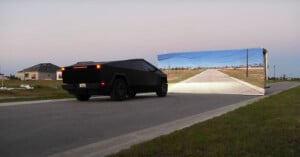 A black, angular vehicle drives toward a realistic painting of a road and landscape on a large vertical poster, positioned across an actual road, creating an optical illusion. The sky is clear, and houses are visible in the background.