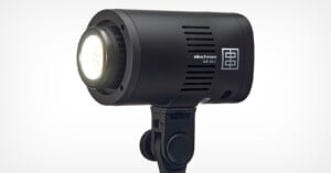 A black Elinchrom LED 100 D light mounted on a stand. The light is turned on, emitting a bright glow. The body has vents for cooling and displays the brand name and model.