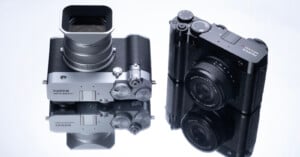 Two Fujifilm cameras are placed on a reflective surface. The camera on the left features a large lens and silver body, while the right camera is more compact with a black finish. Both are mirrored on the surface below them.