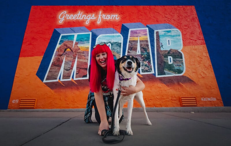 A person with bright red hair smiles while crouching next to a happy dog on a leash. They pose in front of a colorful mural that reads 