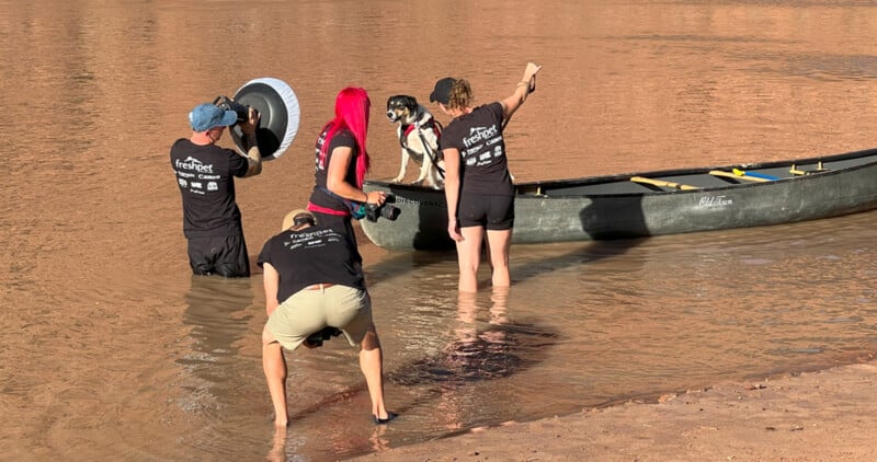 Four people in matching black shirts stand in shallow water, holding a canoe steady. A dog sits inside the canoe. One person points towards the distance, while another holds a large reflective disc. The scene is sunny and involves teamwork.