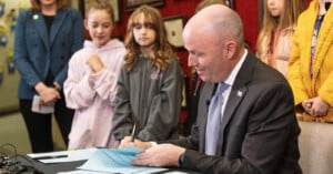 A person in a suit is sitting at a table, signing documents. Several children stand nearby, watching. The background features framed items on a red wall.