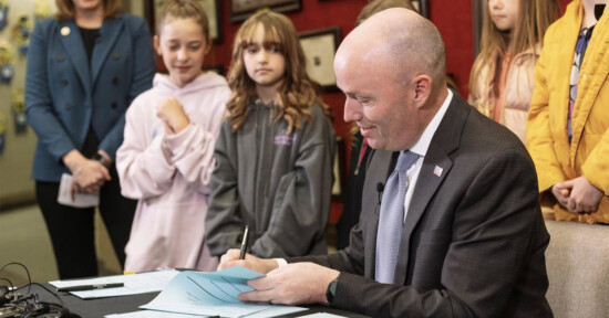 A person in a suit is sitting at a table, signing documents. Several children stand nearby, watching. The background features framed items on a red wall.