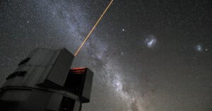 A large observatory telescope emits a bright orange laser into a star-filled night sky. The Milky Way galaxy is visible, with numerous stars scattered across the background.