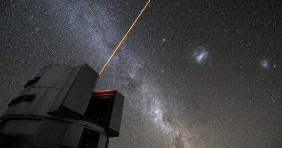A large observatory telescope emits a bright orange laser into a star-filled night sky. The Milky Way galaxy is visible, with numerous stars scattered across the background.