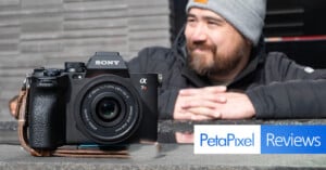 A Sony camera is placed on a stone surface in the foreground, with a leather strap attached. In the background, a person in a grey beanie and black jacket is leaning on the surface, slightly out of focus. The overlay text reads "PetaPixel Reviews.