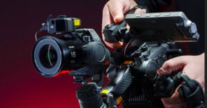 Close-up of a person operating a professional camera setup. The camera features a large lens and a mounted monitor. The background is a dark red, creating an intense atmosphere. The person’s hands are adjusting the camera equipment.