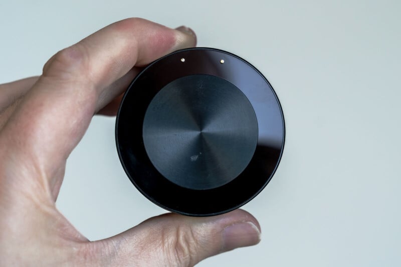 A hand holding a small, round, dark-colored electronic device with two tiny gold contacts on its surface against a plain background. The device has a metallic central circular pattern.