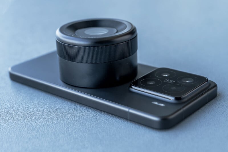 A smartphone with a large, detachable round lens attachment placed on top. The device is resting on a textured blue surface. The phone's camera module is visible beside the lens.