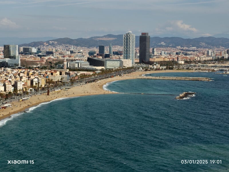 Aerial view of a coastal city with a sandy beach along a wide bay. Skyscrapers and buildings are visible in the background, with mountains in the distance. The ocean is calm, and it's a clear day. The date "03/01/2025" is displayed on the image.