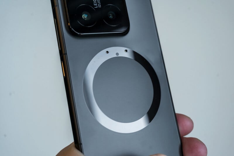 A hand holding a smartphone with a circular magnetic charging area on the back and a dual camera setup near the top. The phone has a sleek, modern design with a metallic finish.