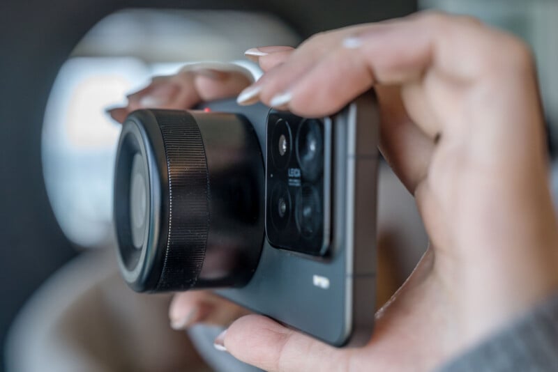 Close-up of a person holding a smartphone with an attached camera lens in both hands. The phone's rear triple camera setup is visible. The background is blurred, highlighting the focus on the device.