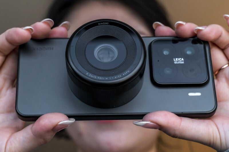 A person holds a smartphone horizontally with both hands. The phone features a large, prominent lens and a Leica-branded camera module on the back. The person's face is blurred in the background.